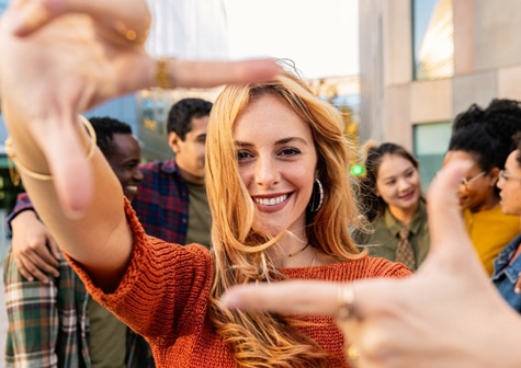 Person posing with hands forming a frame in the foreground, with several people standing outdoors in the background.